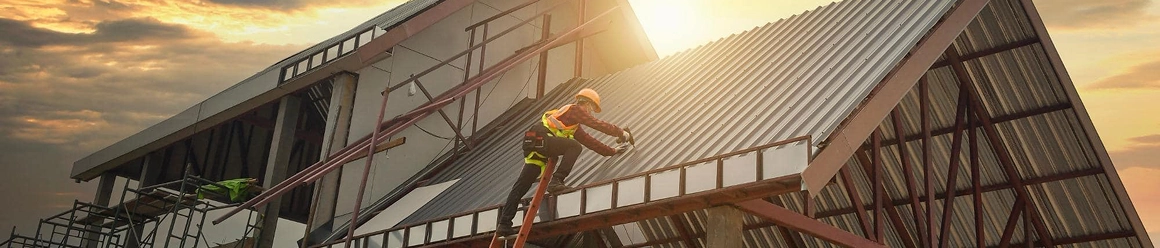 A construction worker in a harness climbing a ladder to install metal siding on the high gable roof of a new building at sunset