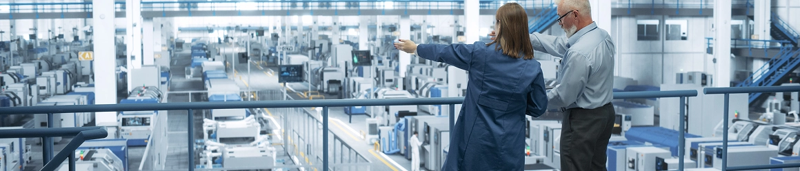 An older man and a young woman standing on a balcony overlooking a vast, brightly lit modern factory floor with machinery
