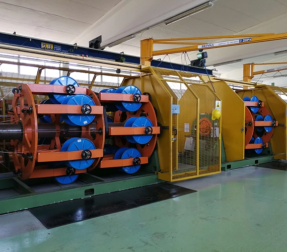 A large, brightly lit industrial machine featuring multiple spools of wire on blue flanges, used for stranding or cabling operations in a clean factory