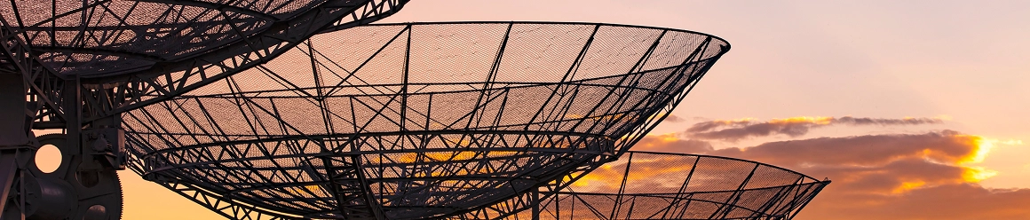 Large, intricate mesh satellite dishes silhouetted against a sky with golden clouds at sunrise or sunset