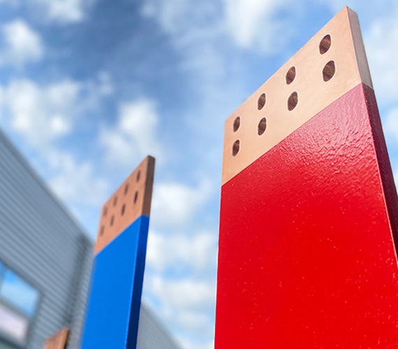 Two large, vertical copper bus bars painted bright red and blue, with exposed copper and bolt holes at the top, set against a blue sky