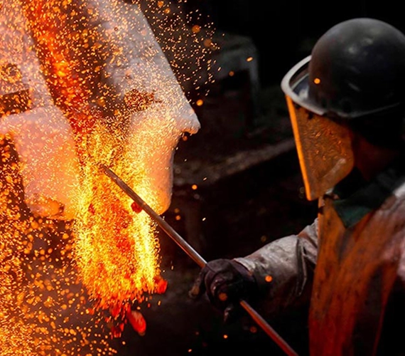A factory worker wearing protective gear and a face shield uses a tool to stir molten metal, causing bright orange and yellow sparks to fly