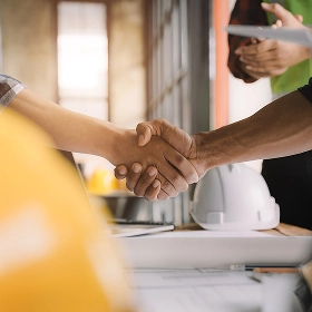 A close-up of two people in a work setting shaking hands over a desk with blueprints and a yellow hard hat
