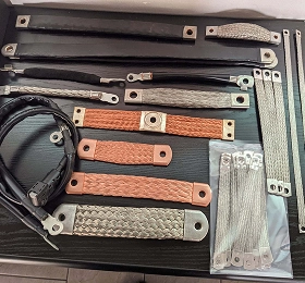 A collection of copper and tin-plated copper flexible connectors, braids, and busbars, including flat and round braided straps with crimped terminals, laid out on a dark surface.